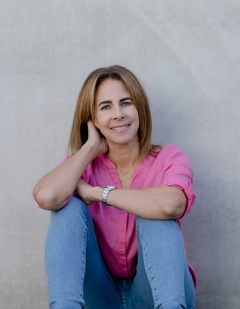 Pamela Klein in Rosa Bluse und Blauen Jeans sitzend vor Betonwand