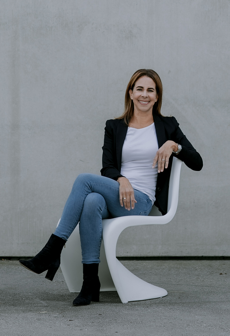 Pamela auf weißem Designer Stuhl sitzend mit weißem T-Shirt blauem Blazer blauen Stiefeln und hellblauer Jeans vor Betonwand